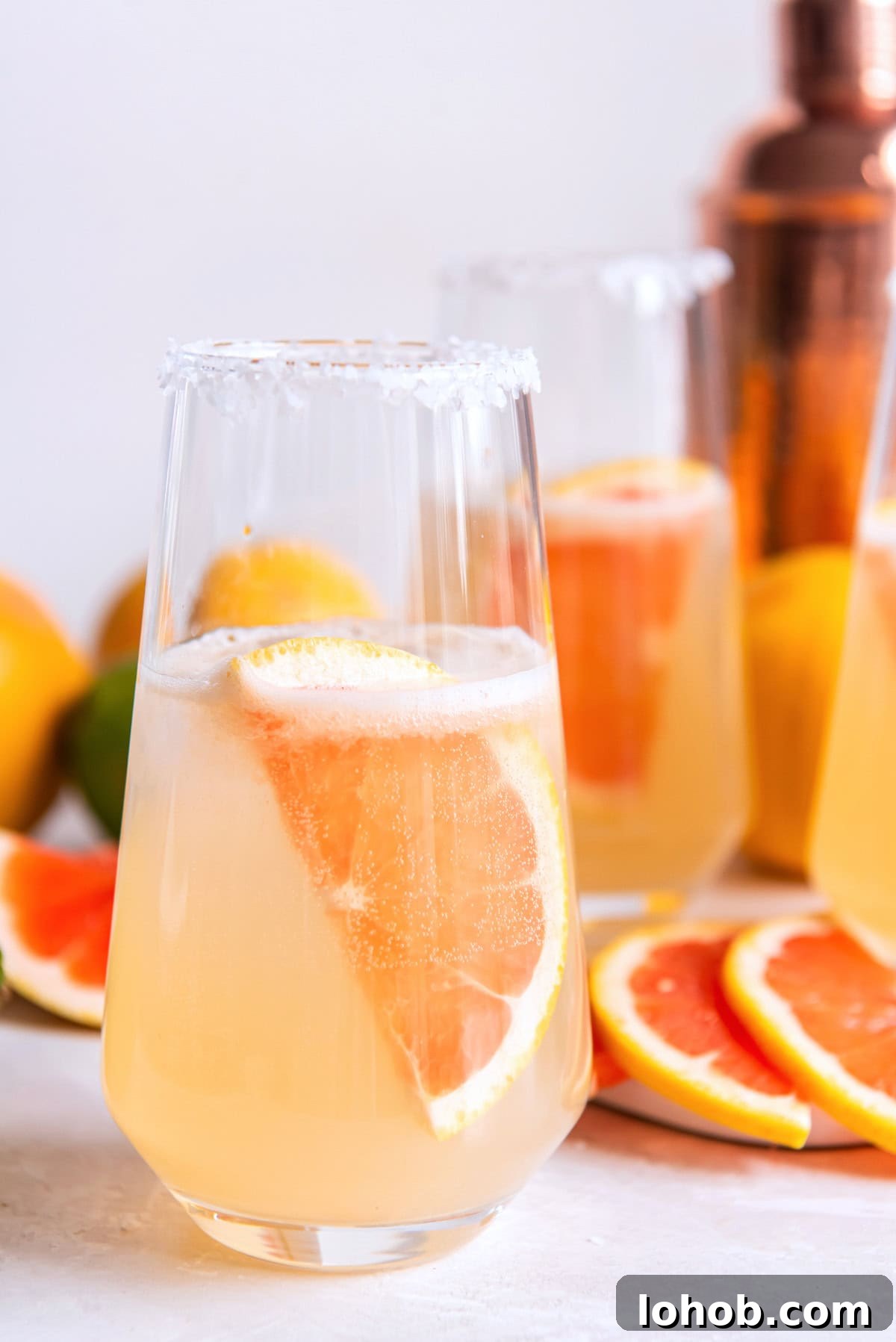 A glass with a pink drink and wedge of grapefruit on a counter with another drink and grapefruits behind it.