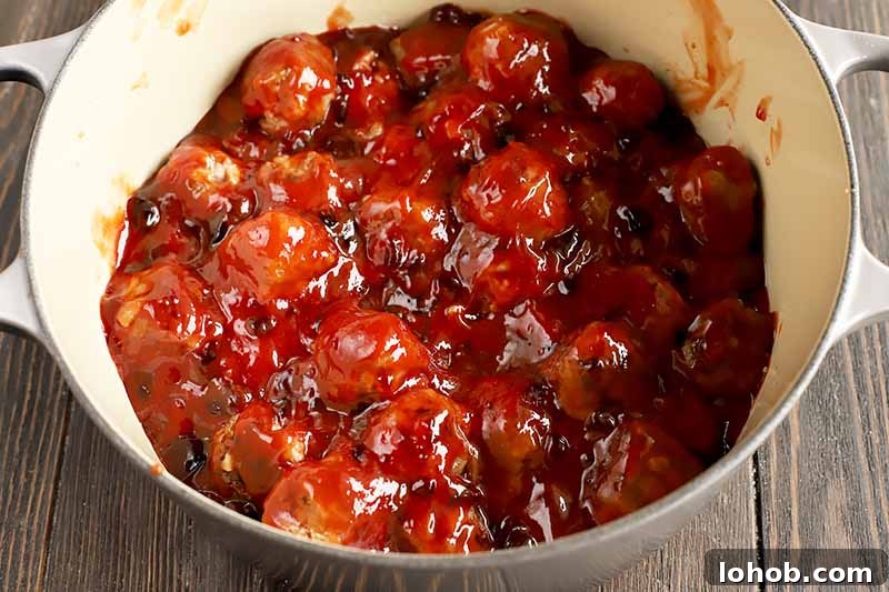 Sweet and Tangy Classic Meatballs Recipe for Weeknight Dinners 13 Horizontal image of meat portions mixed with a thick red liquid mixture in a pot.