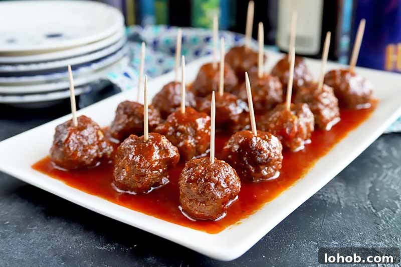 Sweet and Tangy Classic Meatballs Recipe for Weeknight Dinners 14 Horizontal image of a white platter with rows of meatballs covered in a red sauce with toothpicks inserted in them.