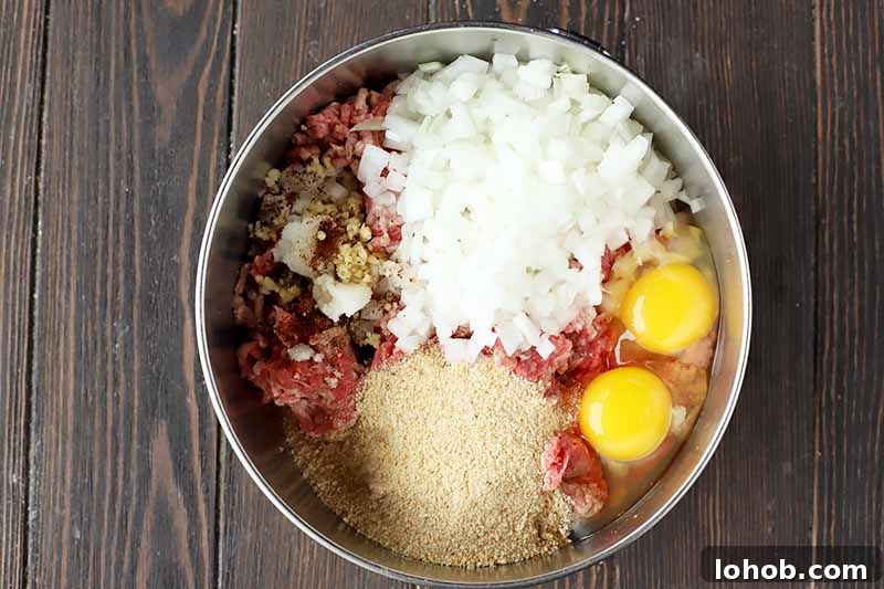 Sweet and Tangy Classic Meatballs Recipe for Weeknight Dinners 8 Horizontal image of eggs, onions, breadcrumbs, and other seasonings on top of raw ground beef in a metal bowl.