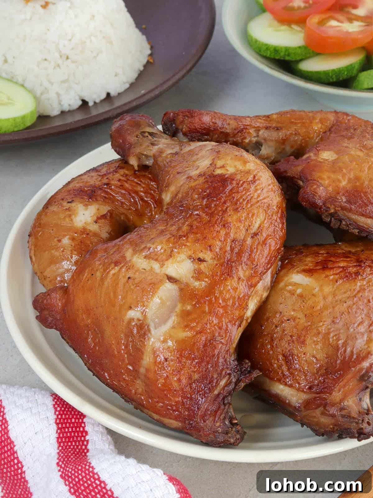 Crispy Soy-Glazed Chicken Recipe for Crunchy, Savory Skin 2 soy sauce chicken on a white platter with rice and sliced cucumbers and tomatoes on the side.