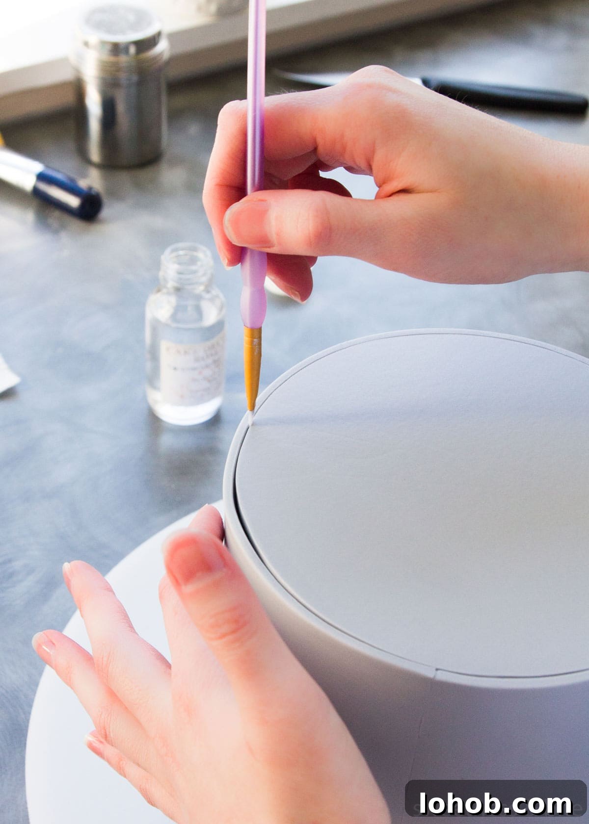 How to Adhere Fondant to Fondant: Guide to Edible Glue 4 A round cake wrapped in grey fondant, with a small paintbrush being used to paint vodka into the join to attach the fondant.