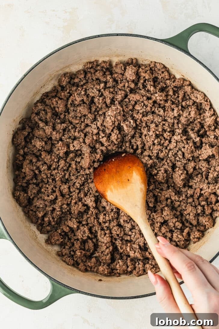 A hand using a wood spoon to stir ground beef in a green Dutch oven.