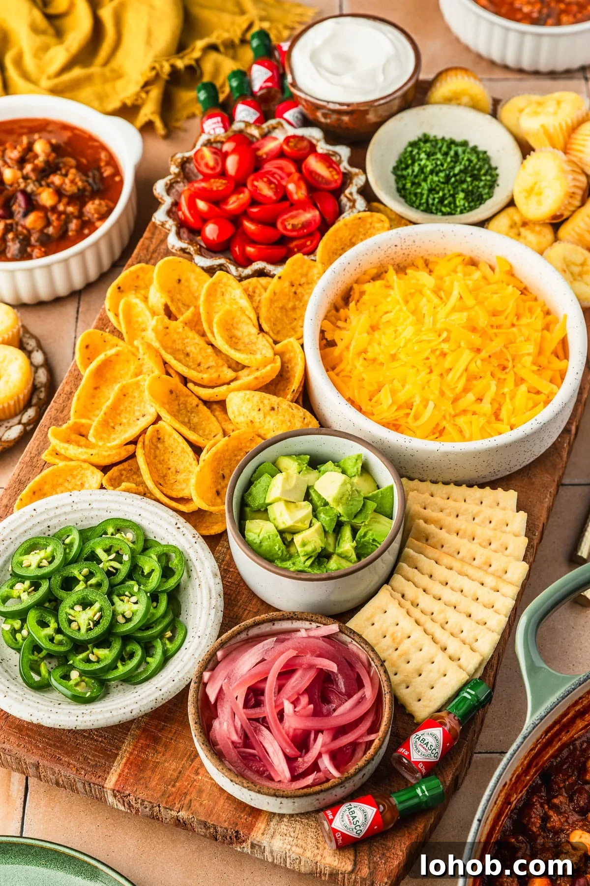 A chili topping charcuterie board with bowls of cheese, veggies, chips, crackers, and herbs on an orange tiled counter next to a pot of chili, bowls of chili, and yellow napkin.