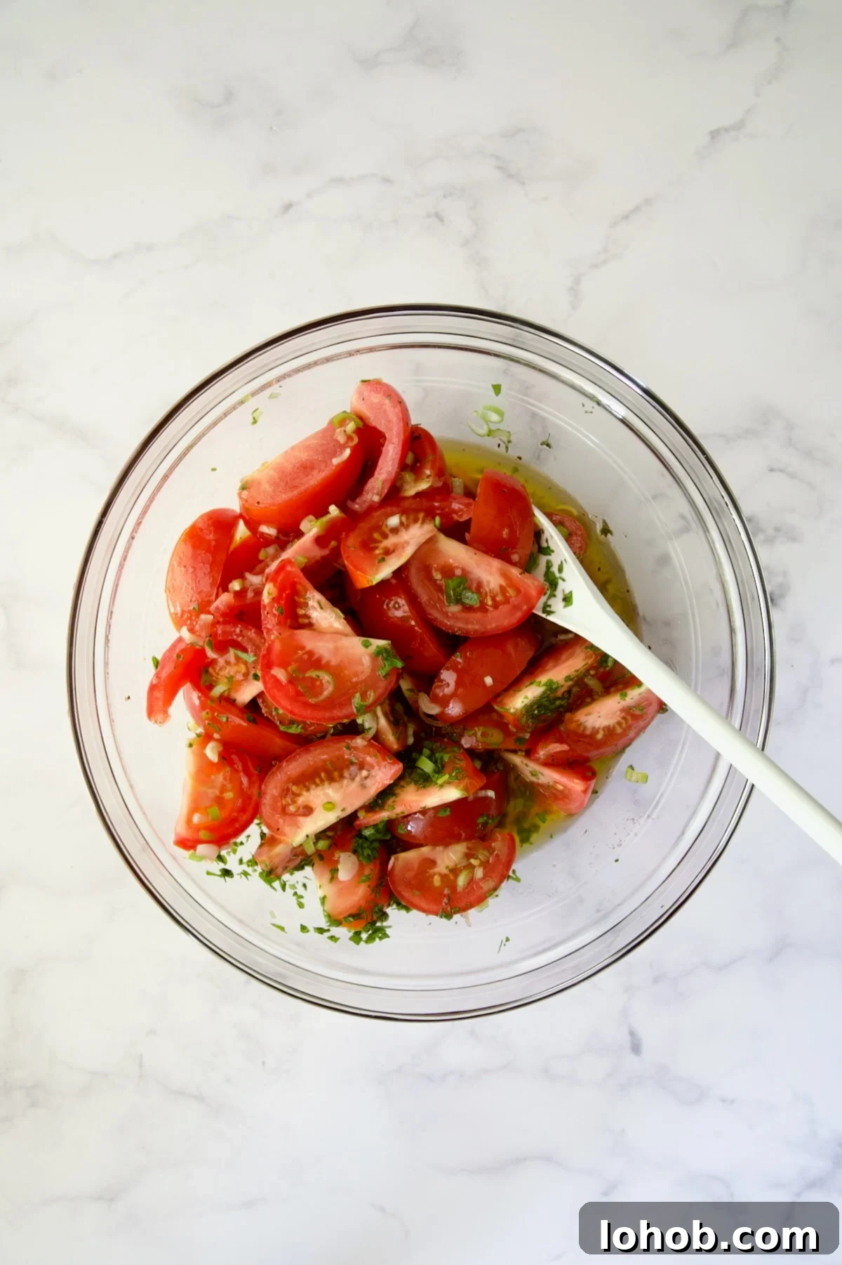 Quick-Pickled Marinated Tomatoes for Fresh Summer Flavor 9 Tomato wedges with dressing, scallions and herbs being stirred in glass bowl with white mixing spoon.