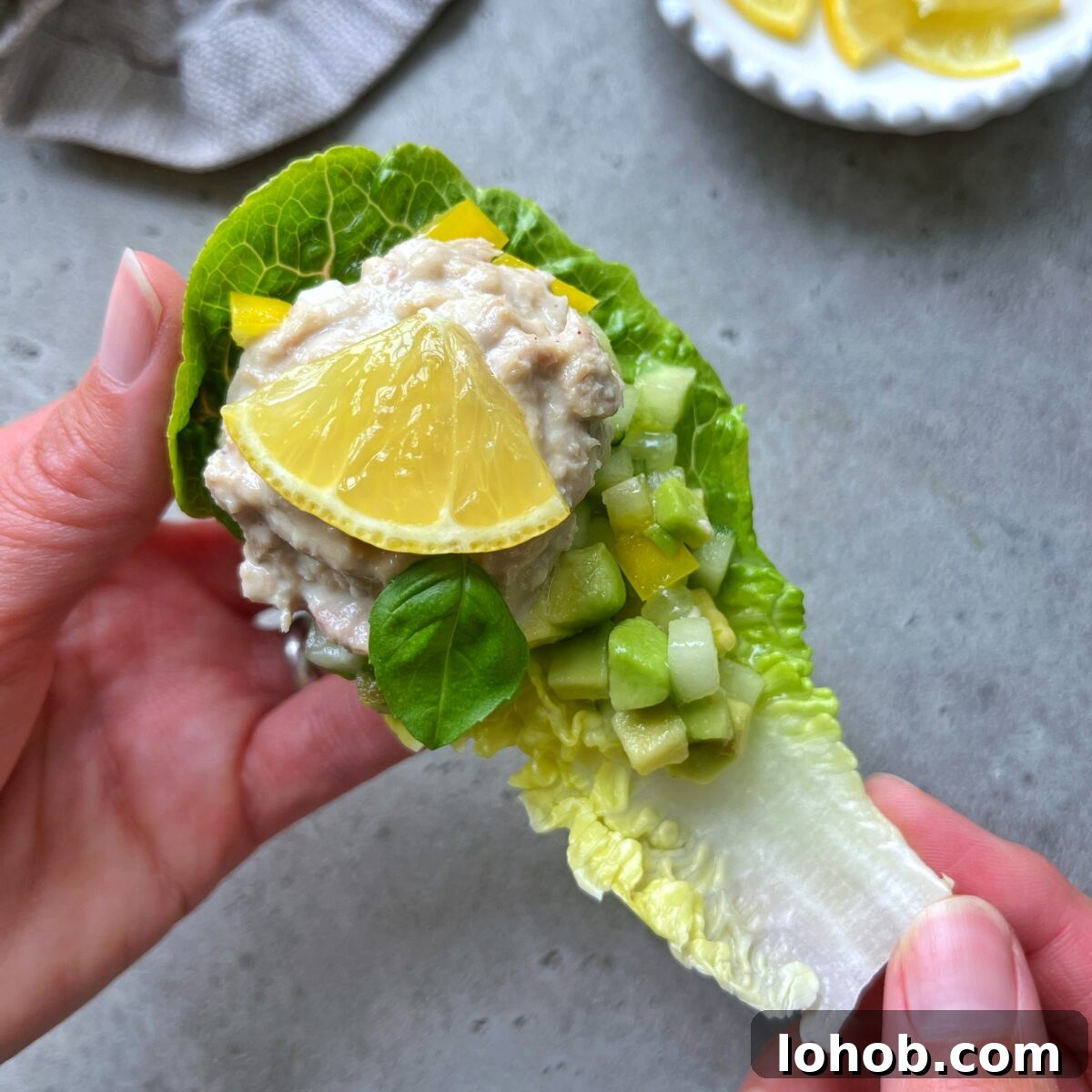Creamy Tuna Salad in Crisp Lettuce Boats 3 A hand holds one of the tuna salad lettuce boats, topped with diced cucumber, yellow bell pepper, a basil leaf, and a lemon wedge.