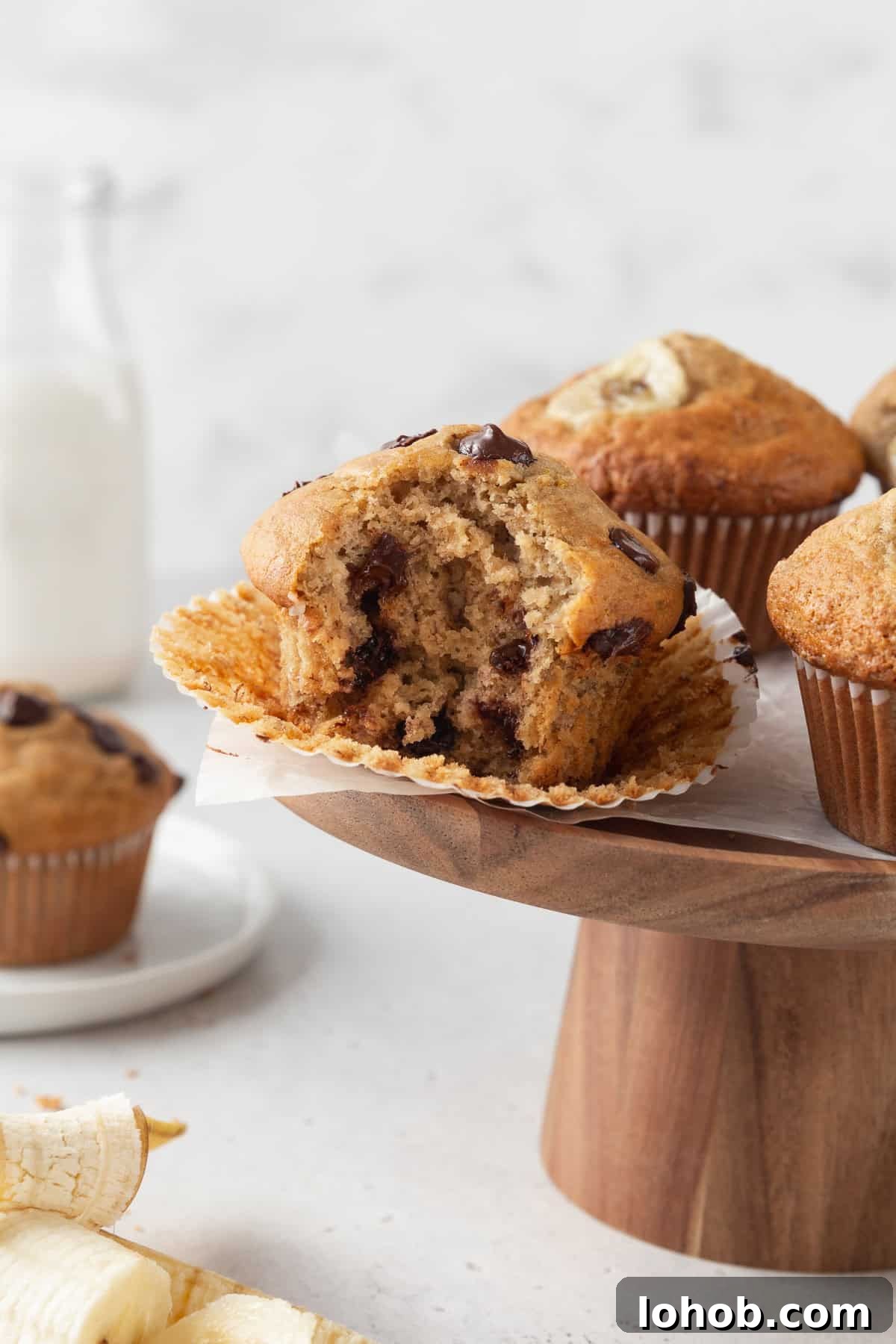 A bitten gluten free banana chocolate chip muffin on a wooden cake stand.