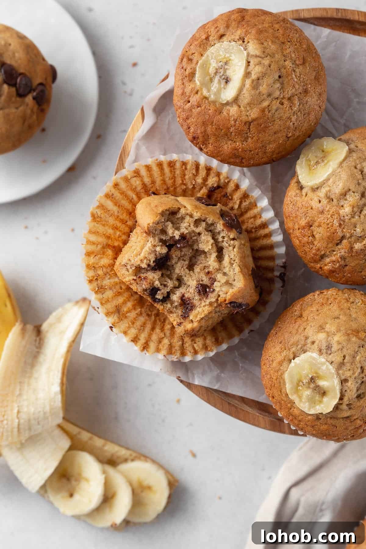 A bitten chocolate chip banana muffin on a paper liner surround by three more muffins.