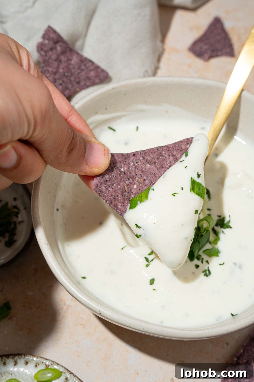 A white bowl with cottage cheese ranch dip garnished with sliced green onion and fresh dill. There is a spoon in the bowl. A hand is dipping a blue corn tortilla chip into the bowl.