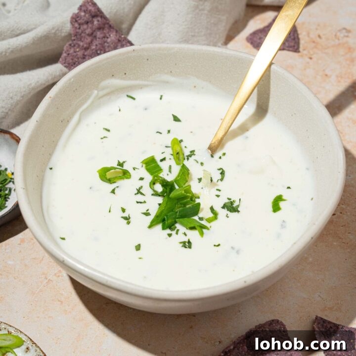 A white bowl with cottage cheese ranch dip garnished with sliced green onion and fresh dill. There is a spoon in the bowl. The bowl is on a tan counter with blue corn tortilla chips.