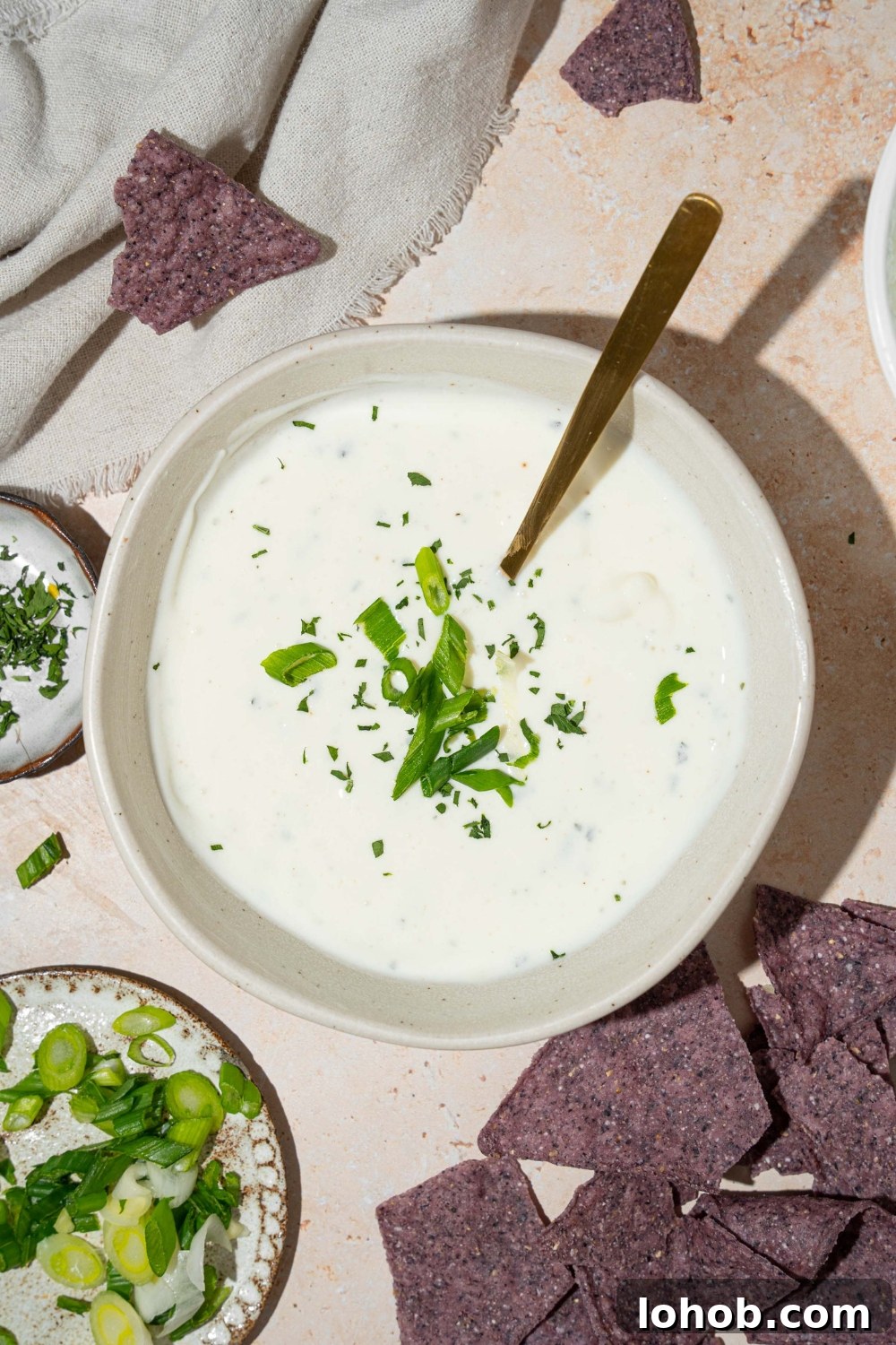 A white bowl with cottage cheese ranch dip garnished with sliced green onion and fresh dill. There is a spoon in the bowl. The bowl is on a tan counter with blue corn tortilla chips.