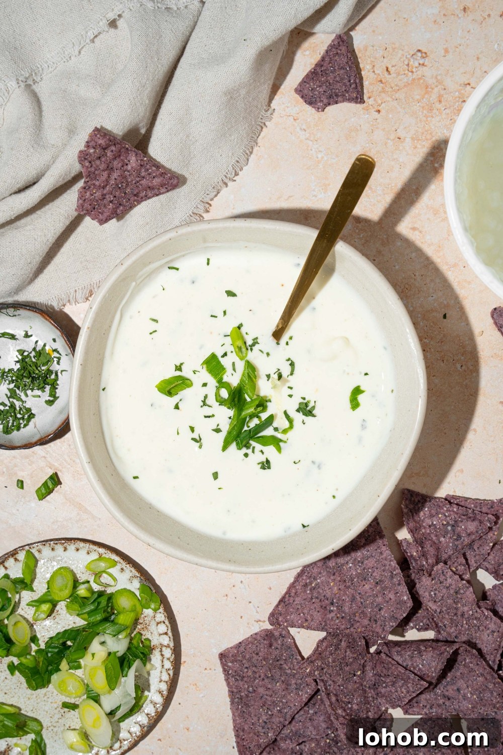 A white bowl with cottage cheese ranch dip garnished with sliced green onion and fresh dill. There is a spoon in the bowl. The bowl is on a tan counter with blue corn tortilla chips.