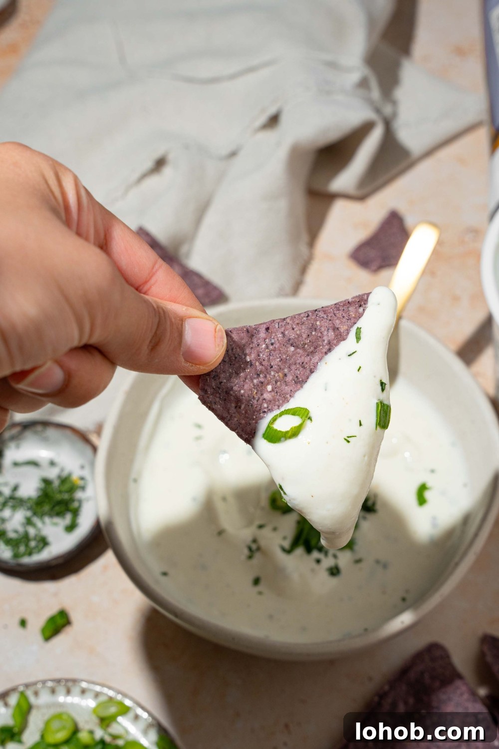 A close up of a hand holding a blue corn tortilla chip dipped in cottage cheese ranch dip. There is a white bowl with cottage cheese ranch dip on a tan counter blurred in the background.