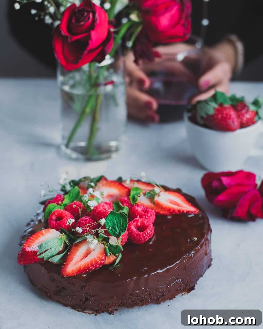 Chocolate cake with berries on white table next to roses and bowl of berries.