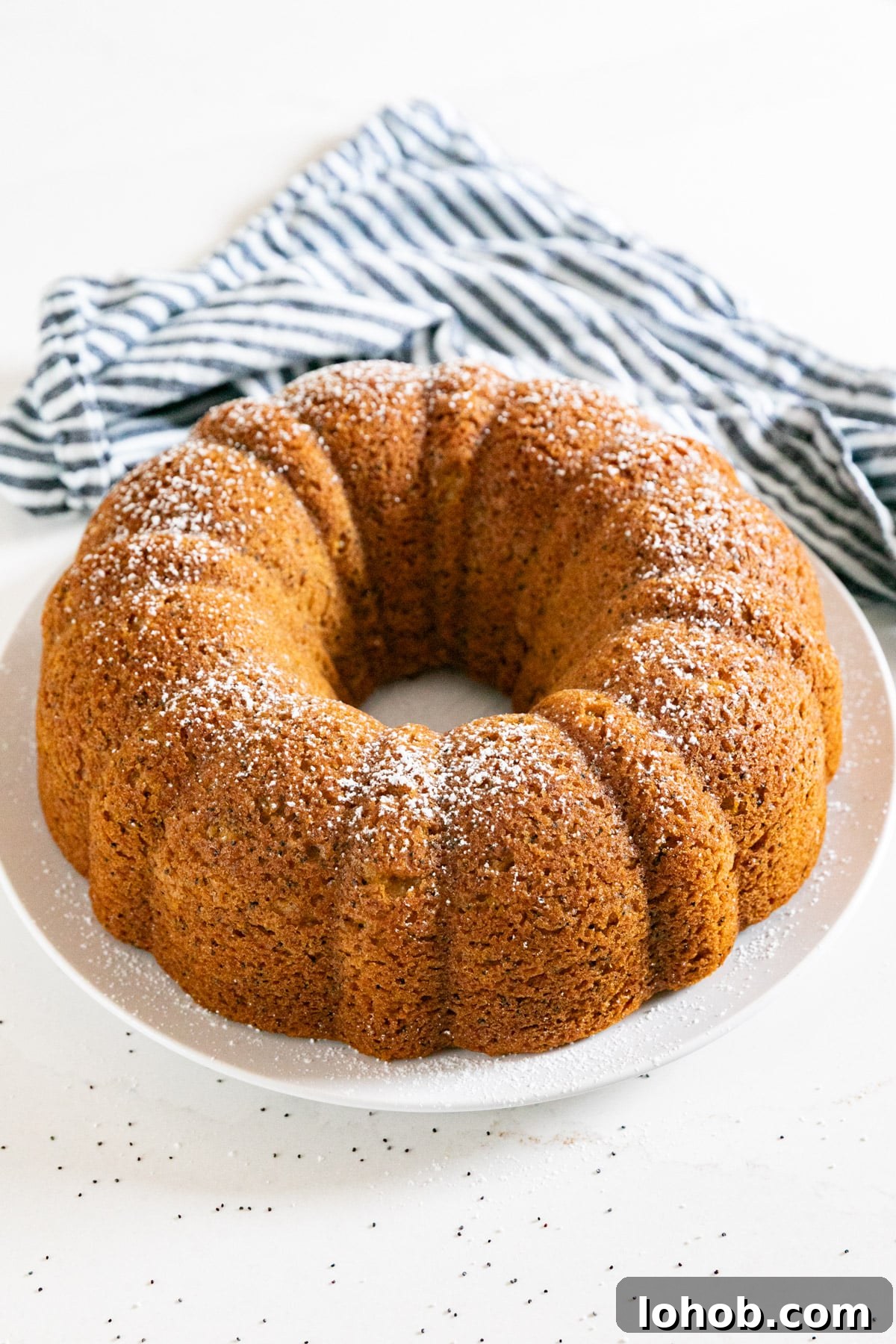 A whole poppy seed bundt cake on a white plate with a striped napkin in the background.