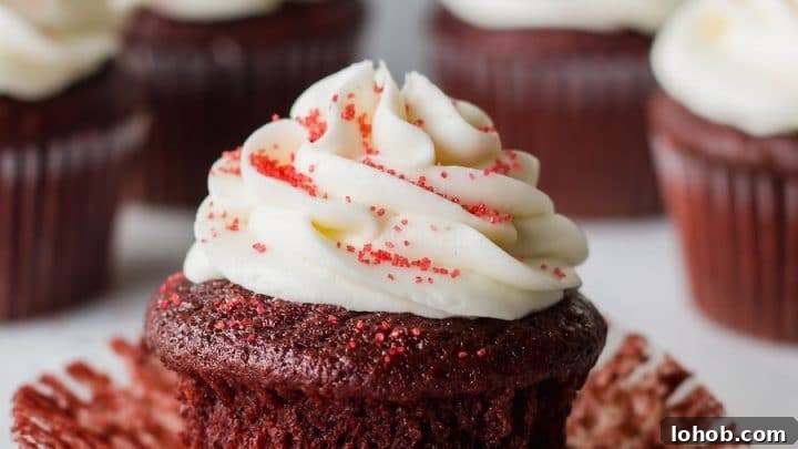 a close up of a gluten free red velvet cupcake topped with white frosting and red sugar, with the paper liner peeled down