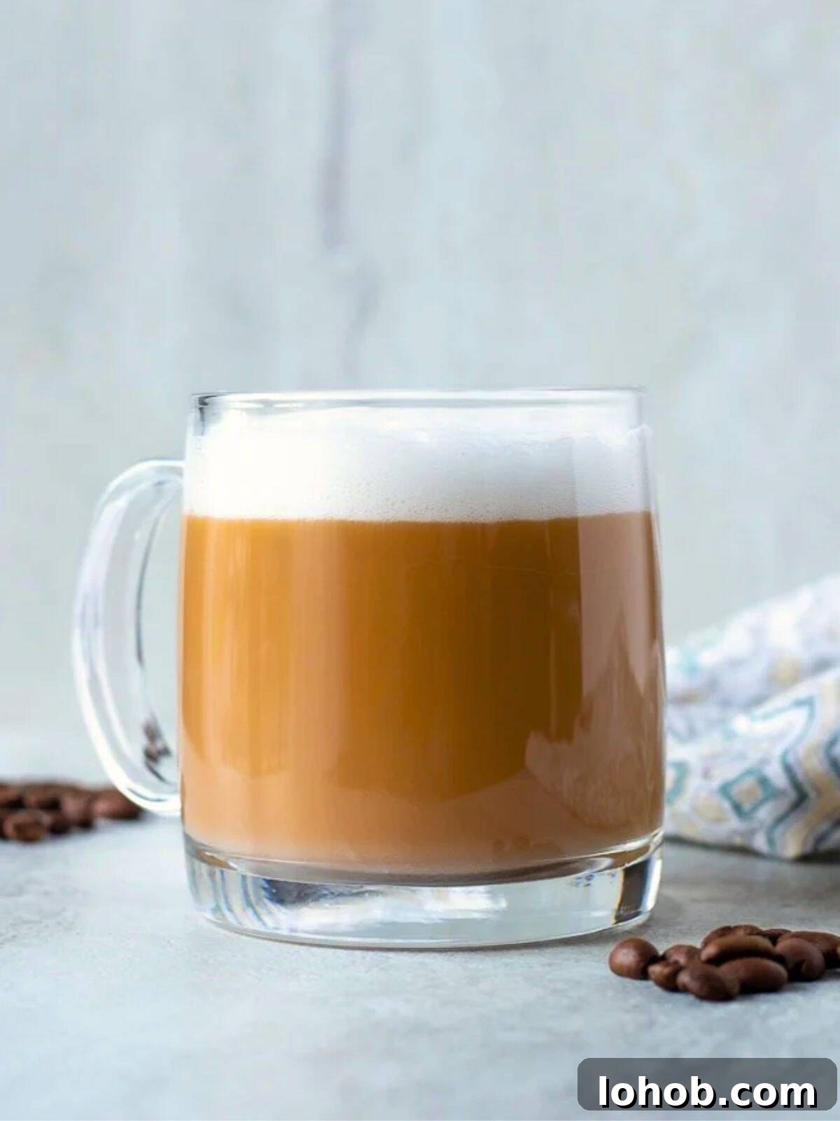 Homemade latte in clear glass coffee mug with thick milk foam and coffee beans in background.
