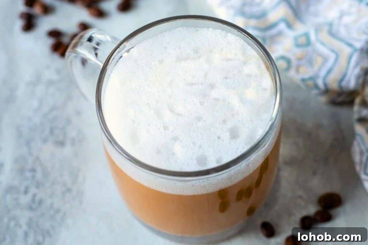 A homemade latte in clear glass coffee mug showing distinct layers of coffee and thick milk foam and coffee beans in background.