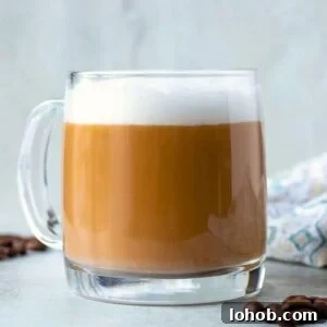 A homemade latte in clear glass coffee mug showing distinct layers of coffee and thick milk foam and coffee beans in background.