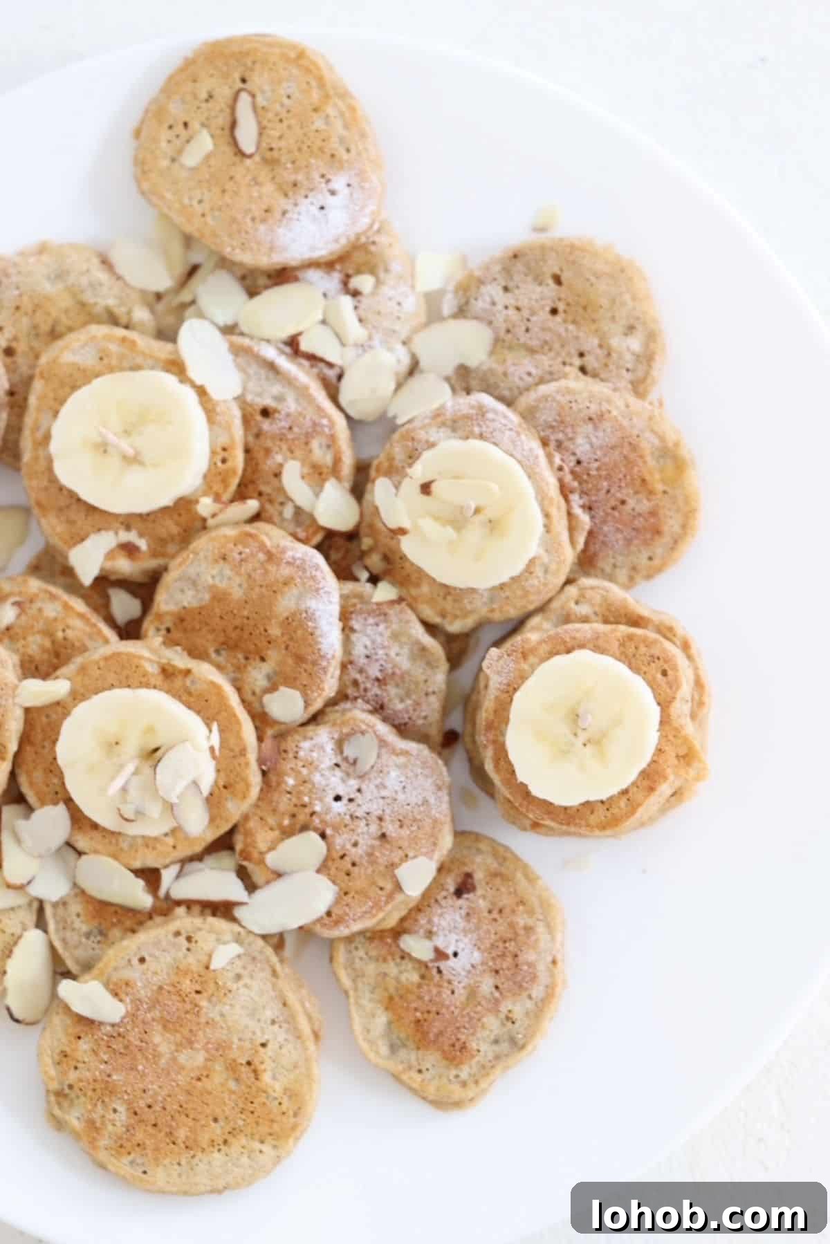 Mini Banana Pancakes Recipe: Fluffy Pancakes with Banana Flavor 2 Mini banana pancakes dusted with powdered sugar, slices of banana, and sliced almond on a white plate.