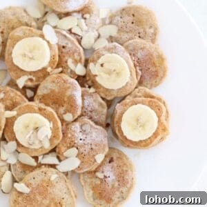 Mini Banana Pancakes Recipe: Fluffy Pancakes with Banana Flavor 9 Mini banana pancakes dusted with powdered sugar, topped with sliced banana and almond slices on a white plate.