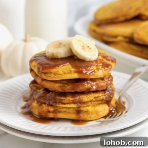 A stack of four banana pumpkin pancakes topped with syrup on a white plate. There are slices of banana on the pancakes and a fork next to them on the plate.