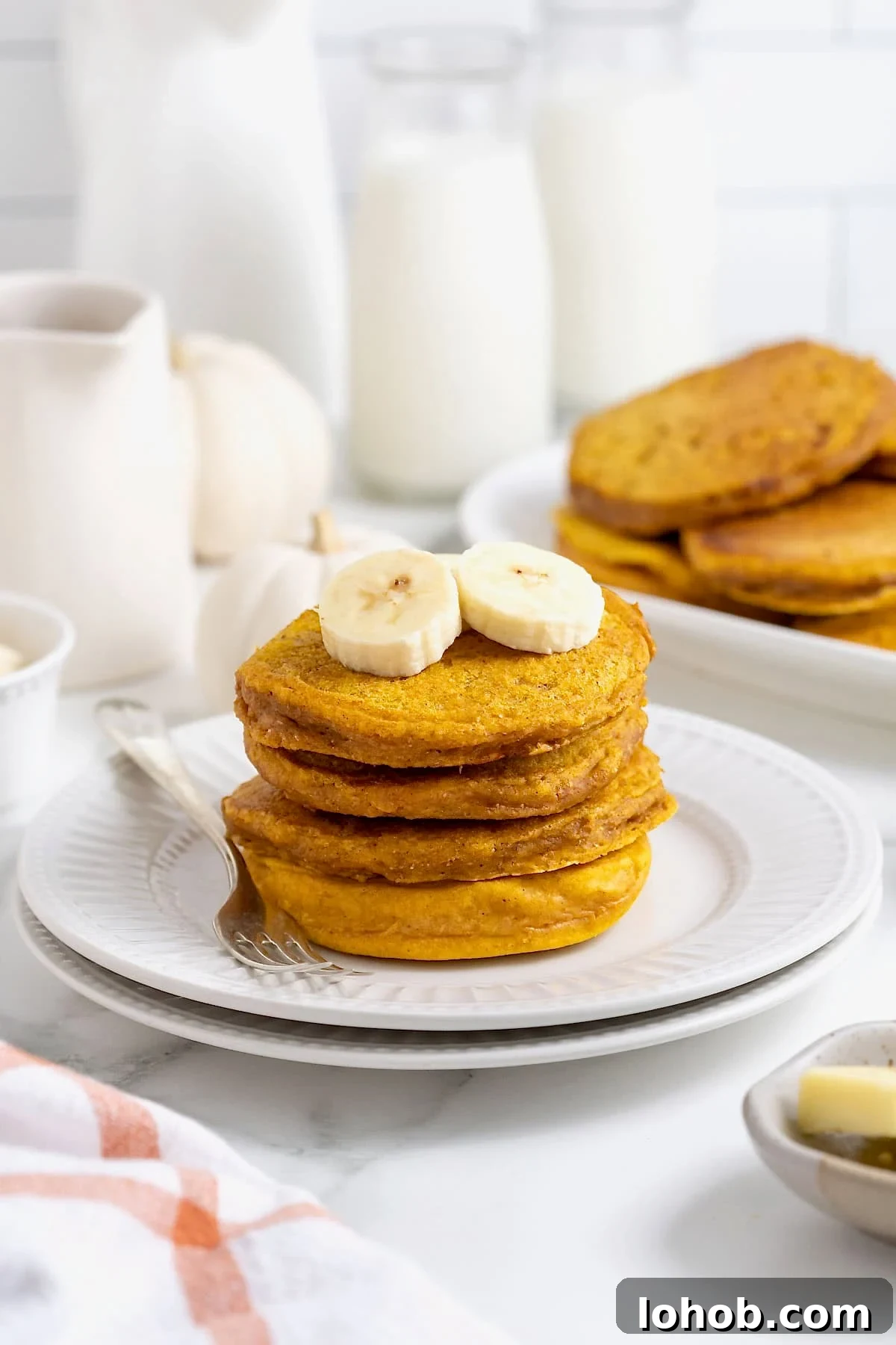 A stack of four pumpkin banana pancakes on a white plate. There are banana slices on top of the pancakes and a fork next to them on the plate.