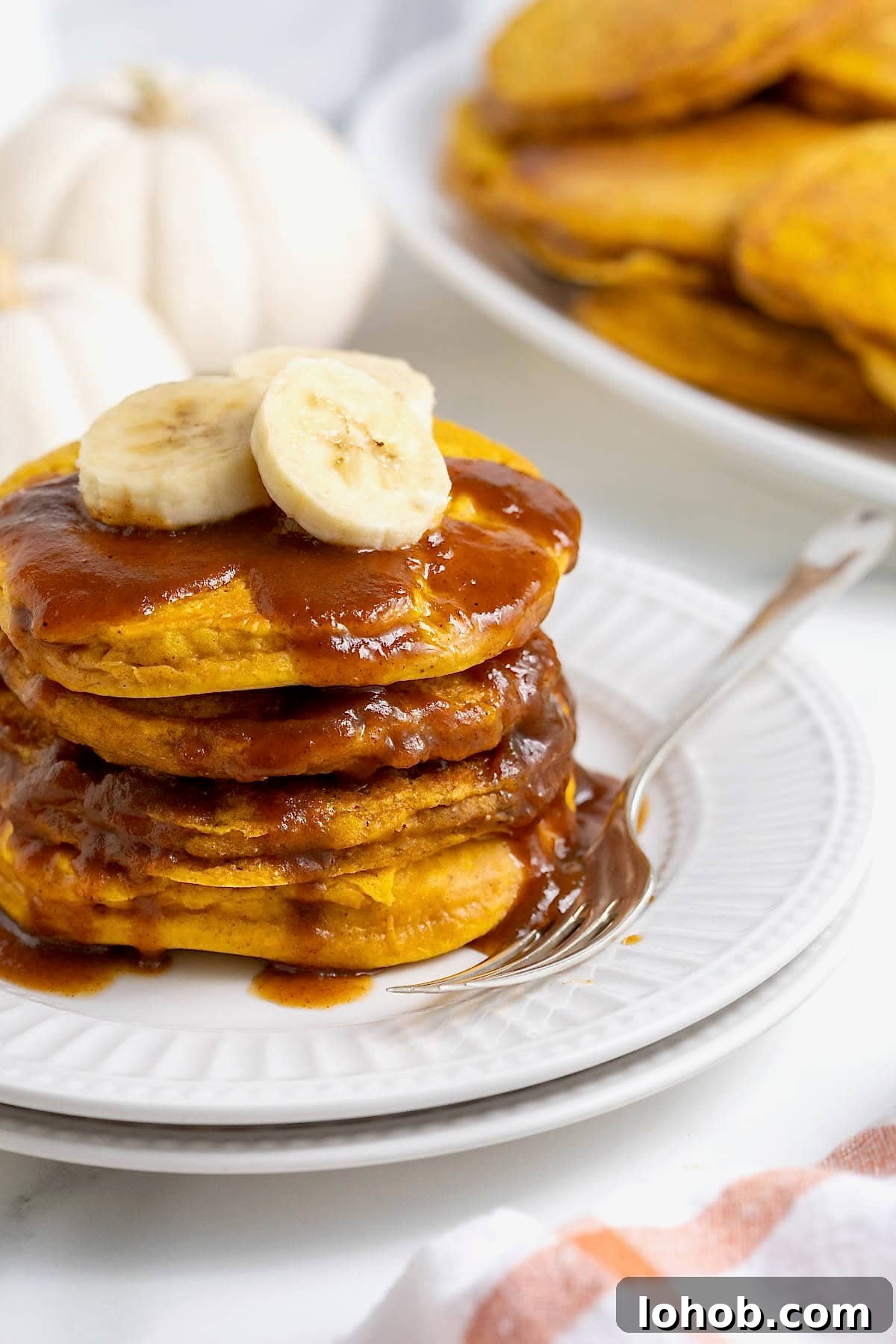 A stack of four banana pumpkin pancakes topped with syrup on a white plate. There are slices of banana on the pancakes and a fork next to them on the plate.