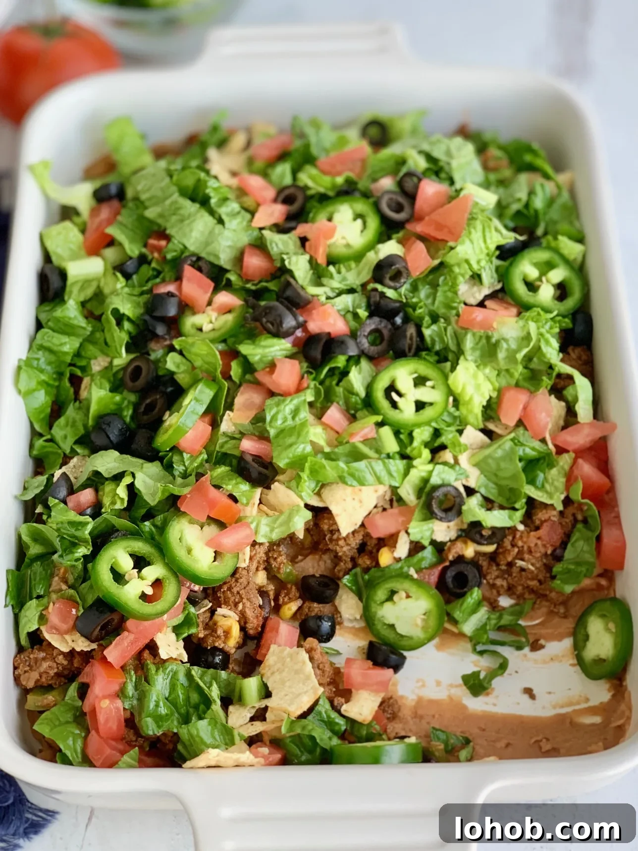 Gluten-Free Dairy-Free Taco Bake Recipe for Weeknight Dinners 8 A baking dish filled with a layer of refried beans, taco meat sauce, crunched up chips, shredded romaine lettuce, diced tomatoes, jalapeno peppers, and black olives.
