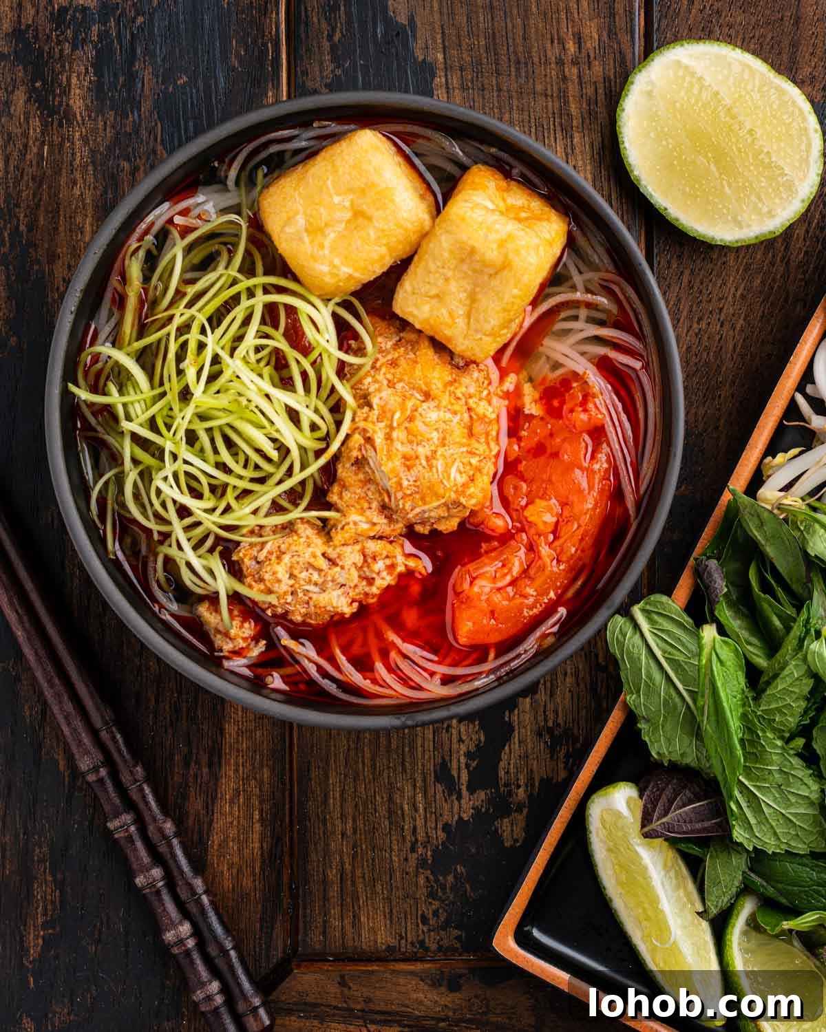 bowl of bun rieu with tomatoes, crab meatballs, water spinach, herbs, and lime.