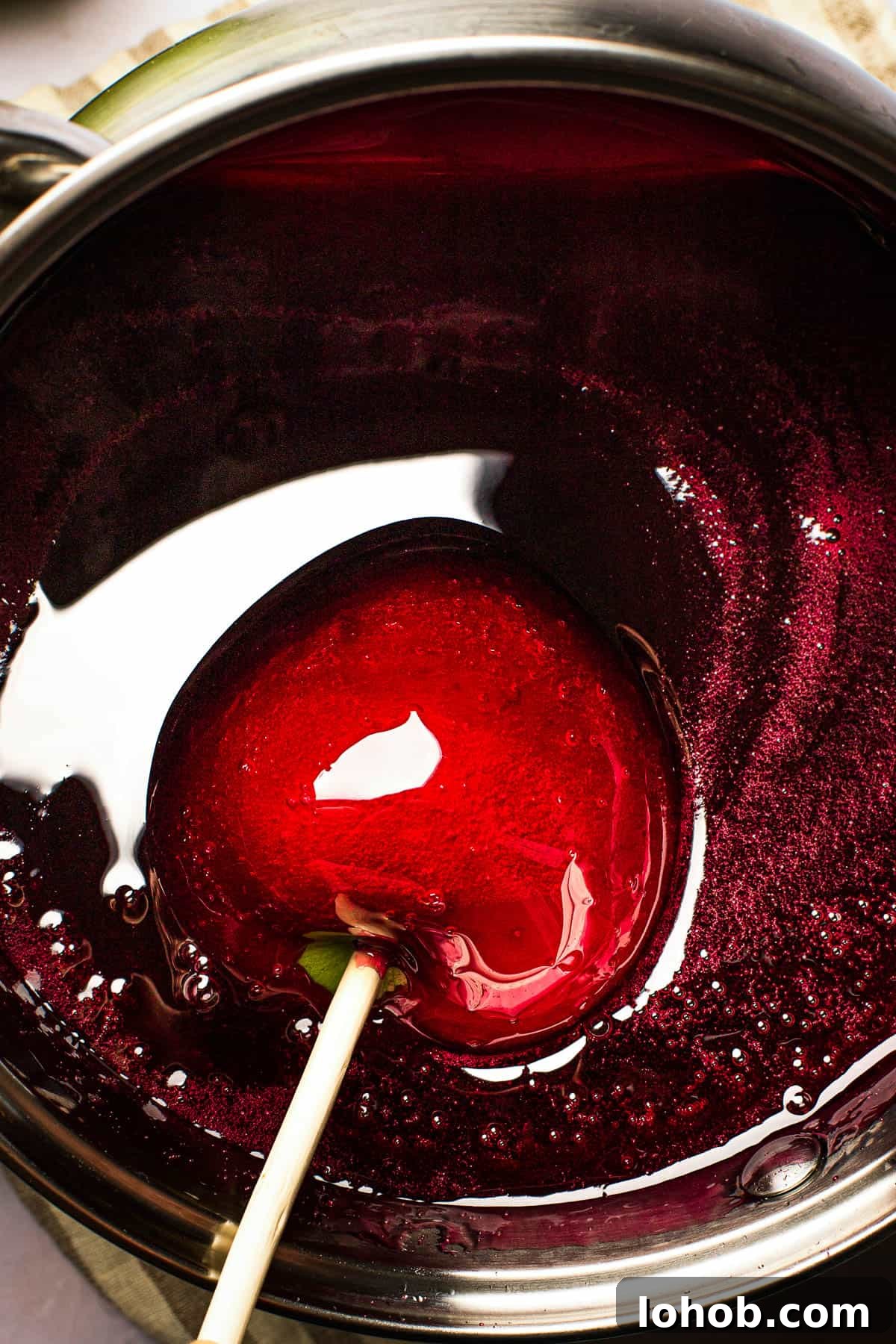 An apple coated in red candy is being lifted from a saucepan of the candy mixture.