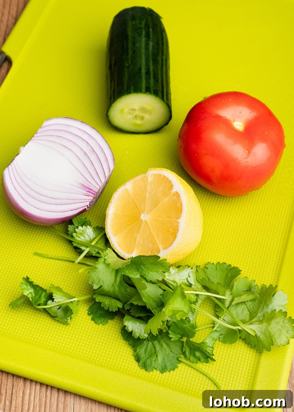 Kachumber: Tangy Indian Onion Salad 4 Quarter of cucumber, a tomato, half a red onion, half of a lemon and fresh coriander on a green chopping board